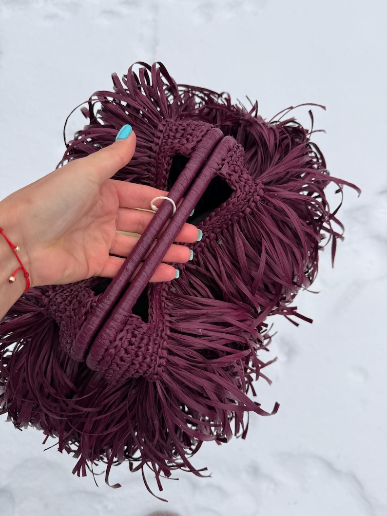 May include: A deep burgundy handbag featuring a dense, layered fringe design covering the entire exterior. The bag is constructed with a crocheted texture and includes two rounded, matching burgundy handles at the top for carrying.