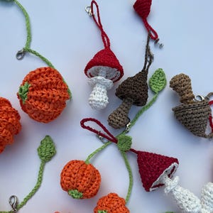 May include: A collection of crocheted ornaments featuring orange pumpkins, red and white mushrooms, and brown mushrooms. Each item includes a green vine with a leaf and a silver metal lobster clasp for hanging, crafted with textured yarn.