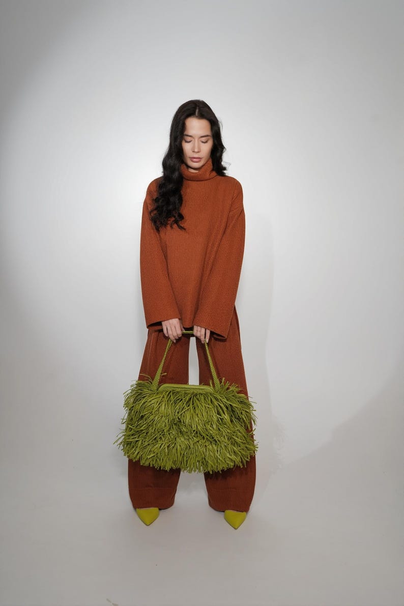 May include: A rust-colored ribbed knit sweater and matching wide-leg trousers paired with a lime green handbag covered in long, dense fringe. The handbag features two top handles and a structured frame, contrasting with the solid knit outfit.