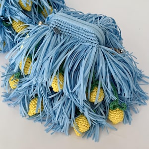Könnte beinhalten: Eine hellblaue Clutch mit einem dichten Fransenbesatz aus dünnen, flachen Streifen. Die Außenseite ist mit mehreren kleinen, gehäkelten gelben Zitronenformen verziert, die an grünen Stielen und Blättern befestigt und in die Fransen eingearbeitet sind.