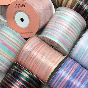 May include: Spools of colorful ribbon in pastel shades of pink, blue, and peach. The ribbon is striped and appears to be made of a woven material. One spool has the brand name "Ispie" printed on the side. The ribbon is likely used for crafts or gift wrapping.