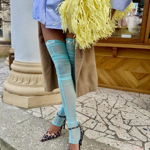 Könnte beinhalten: Transparente, hellblaue Overknee-Strümpfe aus durchscheinendem Netzstoff mit einem kleinen weißen Etikett am oberen Saum. Die Strümpfe werden mit spitzen Schuhen mit Leopardenmuster, einem hellblauen Hemd und einem beigen Mantel kombiniert.