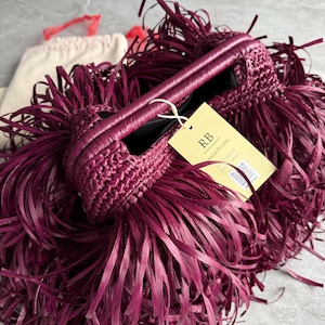 May include: A deep magenta handbag featuring a dense, fringed exterior made of long, thin strips. The bag has a structured, woven top handle and a light yellow rectangular tag attached with a white string, displaying the text RB, special for you.