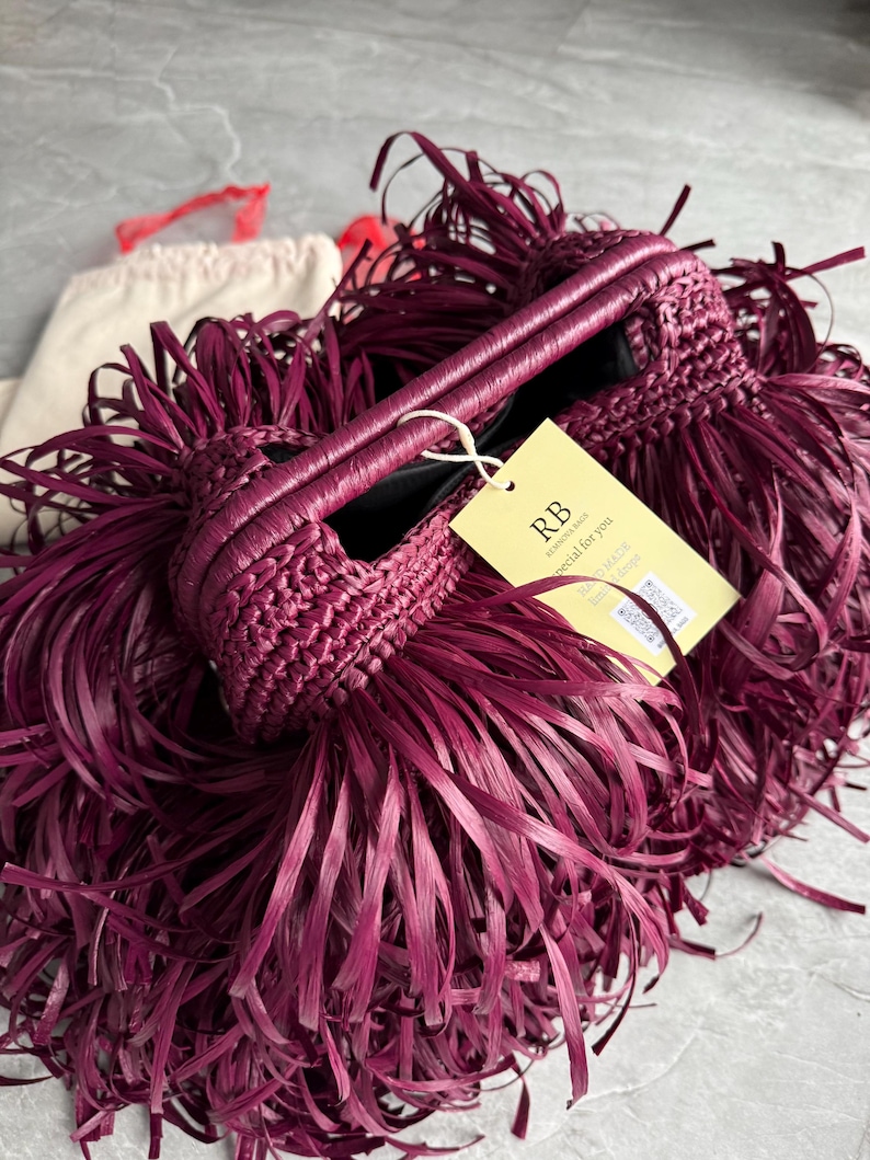 May include: A deep magenta handbag featuring a dense, fringed exterior made of long, thin strips. The bag has a structured, woven top handle and a light yellow rectangular tag attached with a white string, displaying the text RB, special for you.