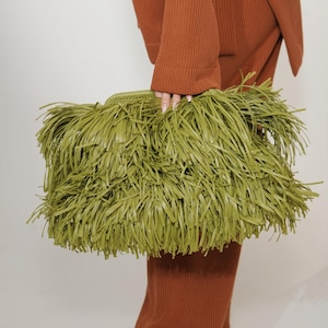 May include: A rectangular olive green clutch bag covered in long, thin, layered fringe strips. The bag features a woven top edge and a structured frame, providing a textured appearance. The material appears to be a smooth, matte synthetic leather.