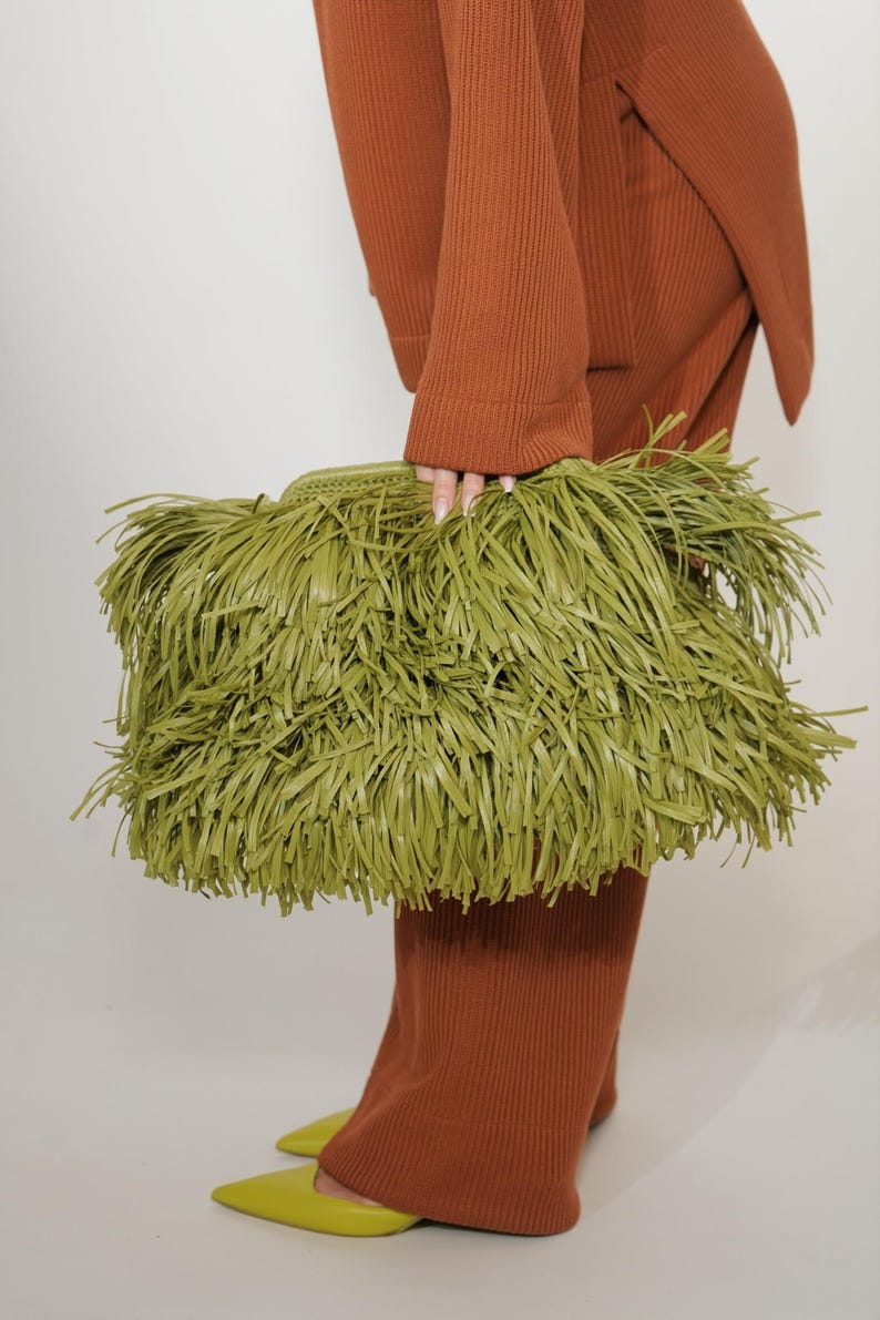 May include: A rectangular olive green clutch bag covered in long, thin, layered fringe strips. The bag features a woven top edge and a structured frame, providing a textured appearance. The material appears to be a smooth, matte synthetic leather.