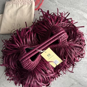 May include: A deep burgundy handbag covered in long, thin, fringed strips with two braided handles. A yellow rectangular tag is attached, featuring the text RB, special for you, and a QR code. A beige fabric dust bag with the logo RB is visible.