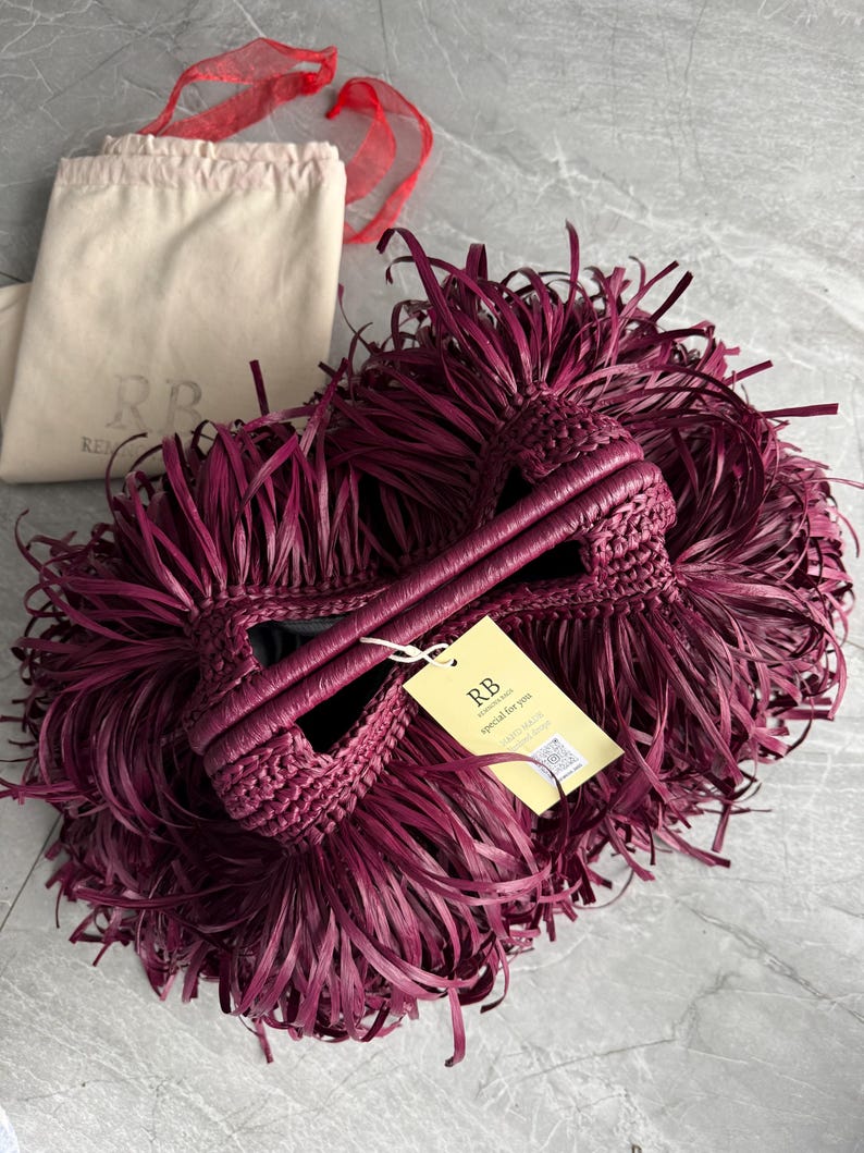 May include: A deep burgundy handbag covered in long, thin, fringed strips with two braided handles. A yellow rectangular tag is attached, featuring the text RB, special for you, and a QR code. A beige fabric dust bag with the logo RB is visible.