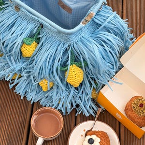 May include: A light blue crochet handbag featuring long fringe and small yellow lemon appliques. The bag has a metal frame closure and a light blue interior lining with a leather tag labeled RB. The bag is open, revealing the interior structure.