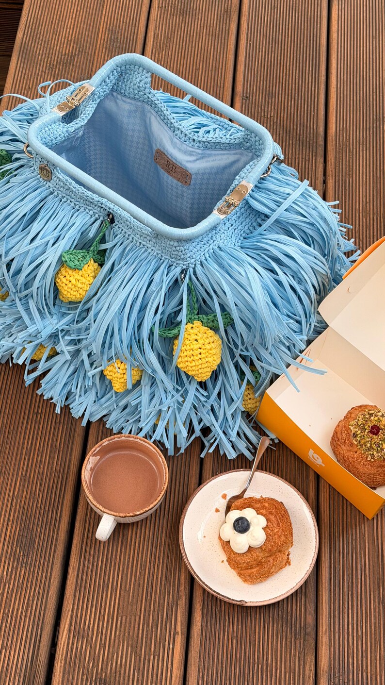 May include: A light blue crochet handbag featuring long fringe and small yellow lemon appliques. The bag has a metal frame closure and a light blue interior lining with a leather tag labeled RB. The bag is open, revealing the interior structure.