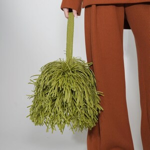 May include: A lime green handbag featuring a dense, shaggy fringe exterior made of thin, hanging strips. The bag includes a single, textured, crocheted-style handle attached to the top, creating a rounded, voluminous silhouette.