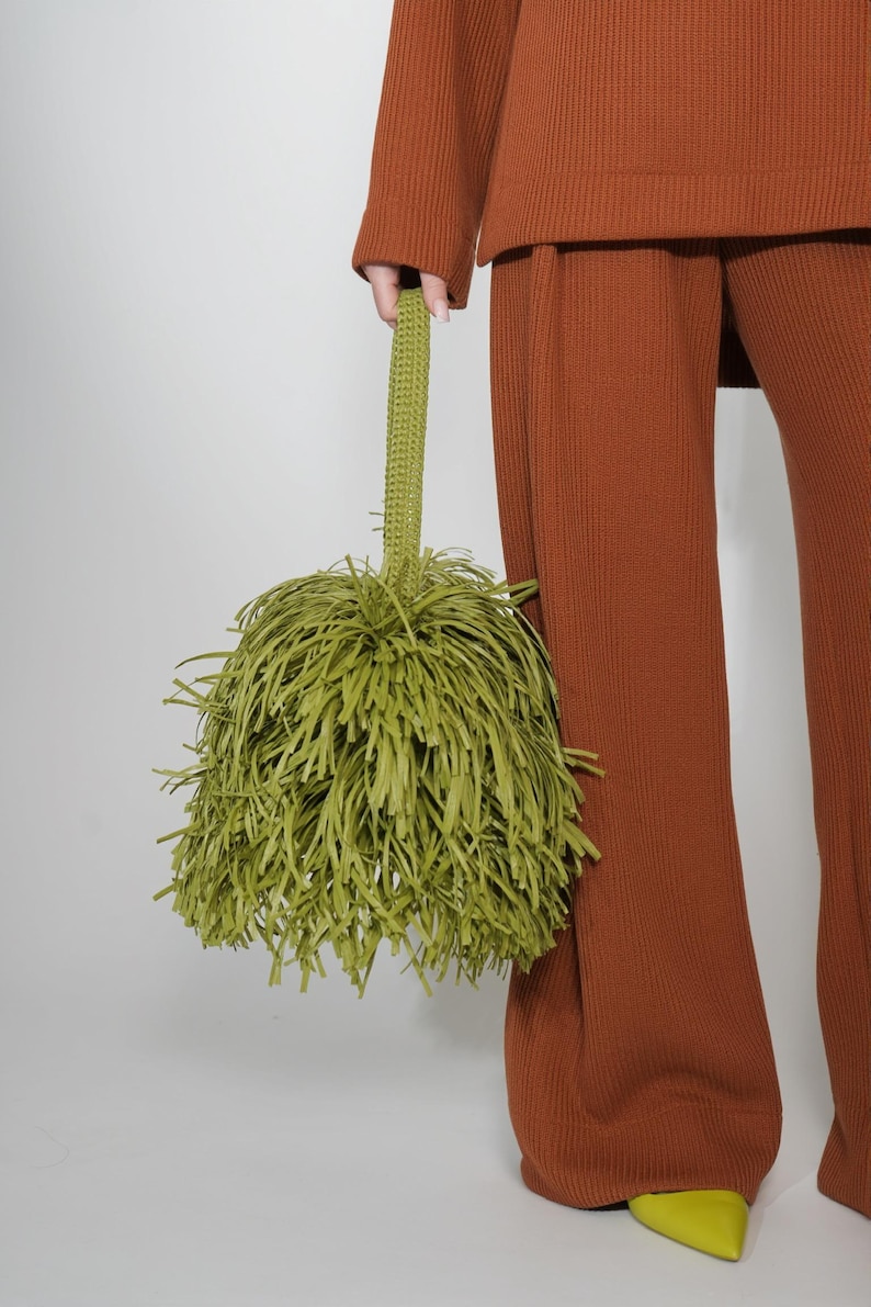 May include: A lime green handbag featuring a dense, shaggy fringe exterior made of thin, hanging strips. The bag includes a single, textured, crocheted-style handle attached to the top, creating a rounded, voluminous silhouette.