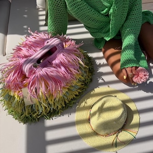 May include: A vibrant handbag covered in layers of pink, yellow, and green fringe, featuring a zippered top closure. Beside it sits a pale yellow straw hat with a decorative band of thin, colourful cords and small beads around the crown.