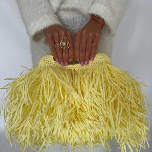 May include: A bright yellow clutch bag featuring a dense, layered fringe design made of thin, flat strips. The bag has a structured, crocheted top opening and a soft, textured appearance throughout, creating a voluminous and layered aesthetic.
