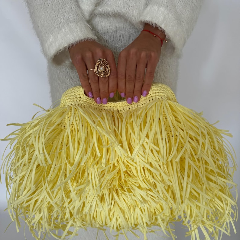 May include: A bright yellow clutch bag featuring a dense, layered fringe design made of thin, flat strips. The bag has a structured, crocheted top opening and a soft, textured appearance throughout, creating a voluminous and layered aesthetic.
