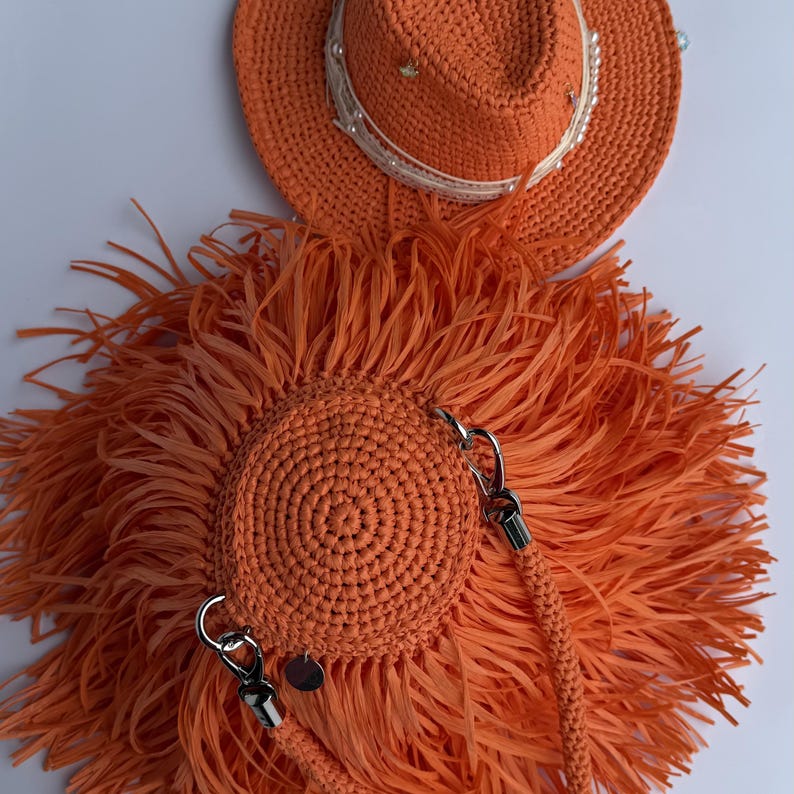 May include: An orange crocheted hat with a white ribbon and pearl embellishments, paired with a matching round orange crocheted bag featuring long fringe, a braided strap with silver-toned metal clasps, and a small circular silver-toned charm.