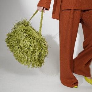 May include: A lime green handbag covered in long, thin, dangling fringe strips. The bag features a woven texture on the top section and two matching green woven handles. The bag is held alongside a rust-colored ribbed long-sleeve top and trousers.