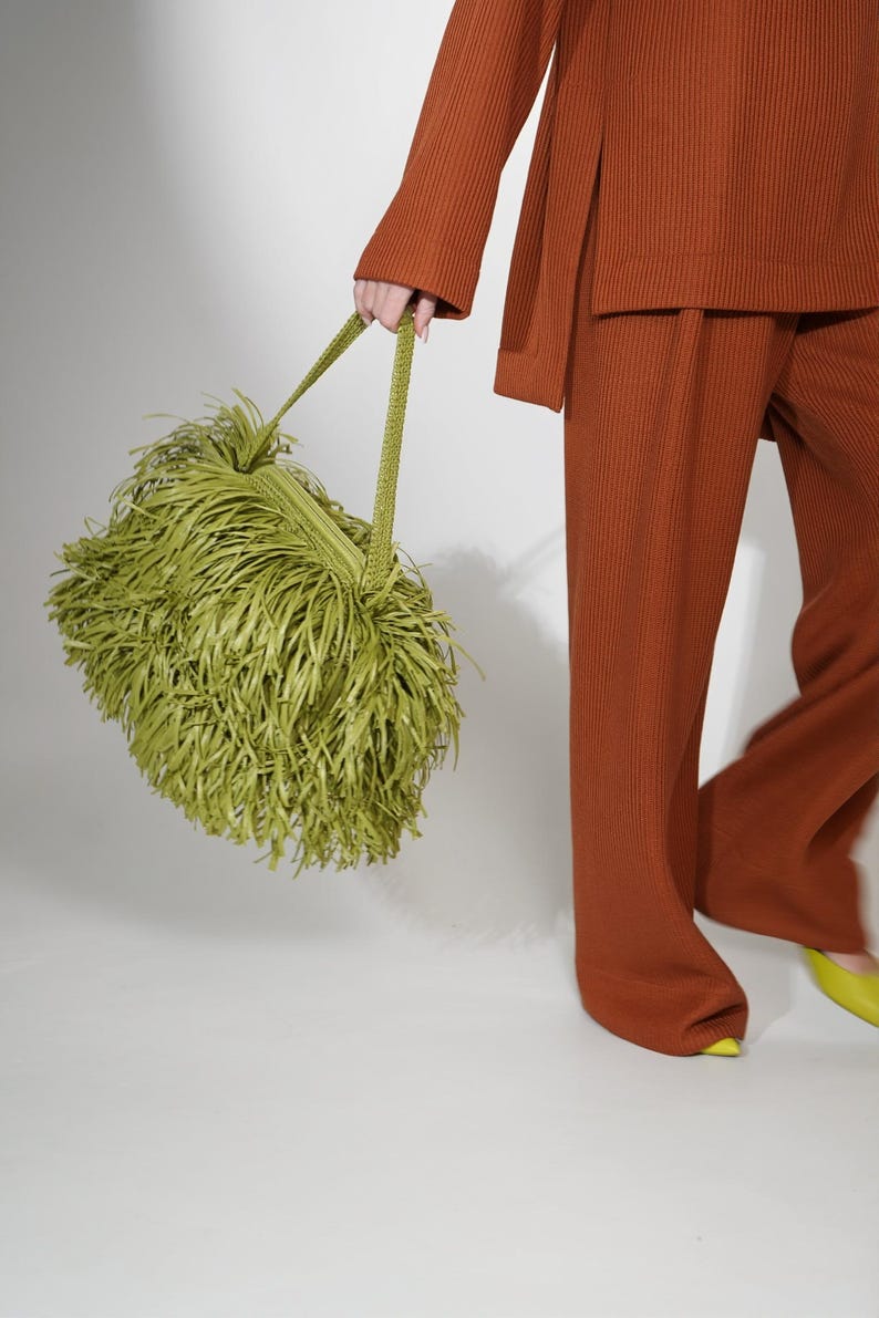 May include: A lime green handbag covered in long, thin, dangling fringe strips. The bag features a woven texture on the top section and two matching green woven handles. The bag is held alongside a rust-colored ribbed long-sleeve top and trousers.