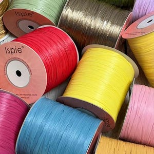 May include: A collection of colorful spools of raffia ribbon in various shades, including red, yellow, blue, pink, and green. The spools are labeled with the brand name "Ispie". Ideal for crafting and gift wrapping.
