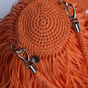 May include: A bright orange crocheted bag featuring a circular, tightly stitched center panel surrounded by long, flowing fringe. The bag includes silver-toned metal lobster claw clasps and a small circular metal tag attached to the side hardware.