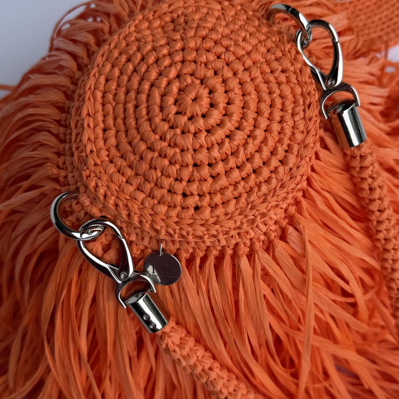 May include: A bright orange crocheted bag featuring a circular, tightly stitched center panel surrounded by long, flowing fringe. The bag includes silver-toned metal lobster claw clasps and a small circular metal tag attached to the side hardware.
