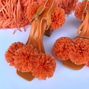 May include: Orange high-heeled sandals with a square toe and ankle strap. The sandals feature large, fluffy orange pom-poms on the toe strap and ankle strap. The heels are a block style, and the sandals are made of a leather-like material.