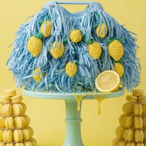 May include: A light blue crocheted handbag featuring a fringe texture and attached yellow crocheted lemon motifs with green leaves. The bag sits on a light green pedestal stand, flanked by two towers of yellow macarons and fresh lemon slices.