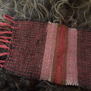 Peut inclure: Un textile rectangulaire tissé à la main présentant un motif rayé dans des tons de brun foncé, de rouge rouille et de rose sourd. Le tissu a une texture tissée grossière visible et comprend une série de franges en fil rouge sur un côté.