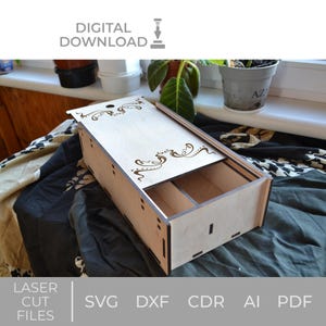 Könnte beinhalten: Eine Holzkiste mit einem Schiebedeckel und zwei Fächern. Die Kiste ist mit einem Laser-geschnittenen Design aus wirbelnden Linien und einem kleinen Kreis verziert. Die Kiste ist aus hellem braunem Holz gefertigt und hat eine natürliche Oberfläche. Die Kiste steht auf einem schwarz-weiß gemusterten Stoff.