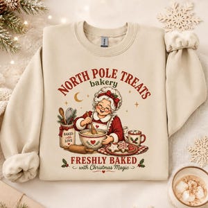 May include: A beige crewneck sweatshirt featuring a graphic of a woman with glasses and a white apron mixing dough in a bowl. The text reads NORTH POLE TREATS bakery FRESHLY BAKED with Christmas Magic. The design includes cookies and a mug.
