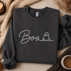 May include: A dark gray crewneck sweatshirt featuring white embroidered text that reads Boo, with the letter o replaced by a simple line-drawn ghost figure. The garment has long sleeves with ribbed cuffs and a ribbed neckline, showcasing a clean design.