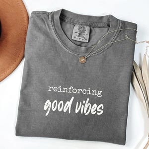 Peut inclure: Un t-shirt gris Comfort Colors avec les mots "reinforcing good vibes" en écriture blanche. Un collier en or avec un pendentif repose sur le t-shirt. Un chapeau marron et une plante séchée sont en arrière-plan.