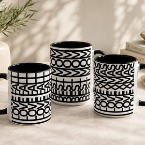 May include: Three ceramic mugs with black interiors and handles. The white exteriors feature black graphic patterns. One mug displays the repeating text LOVE ALL, another shows the text BEST MOM, and the third features the repeating text LOVE XMAS.