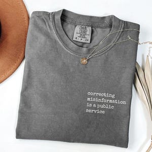 May include: A gray Comfort Colors t-shirt with the text "correcting misinformation is a public service" in white. A silver necklace with a small pendant rests on the shirt. A brown hat is visible in the background.
