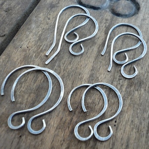 May include: Five silver-toned metal earring hooks. The hooks are all in the shape of a loop with a small hook at the end. The hooks are arranged in a circle on a wooden surface.