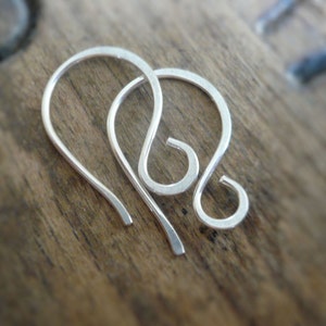 May include: Two silver earring hooks, made of wire, are shown on a wooden surface. The hooks are in the shape of a teardrop with a small loop at the top.