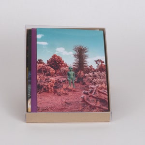 Pode incluir: Um conjunto de três livros com uma lombada roxa e uma imagem de capa de uma paisagem desértica com uma figura verde em pé no meio.