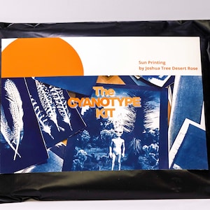 Pode incluir: Uma embalagem preta contendo um kit de cianotipia. A capa do kit apresenta um gráfico de sol laranja e o texto "Sun Printing by Joshua Tree Desert Rose". No interior, imagens azuis e brancas são visíveis, com as palavras "The Cyanotype Kit."