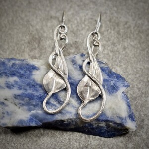 May include: A pair of silver-toned drop earrings featuring a stylized leaf design with a curved, swirling metal wire frame. The earrings have a polished finish and are suspended from simple hook-style ear wires, showcasing a delicate, organic shape.