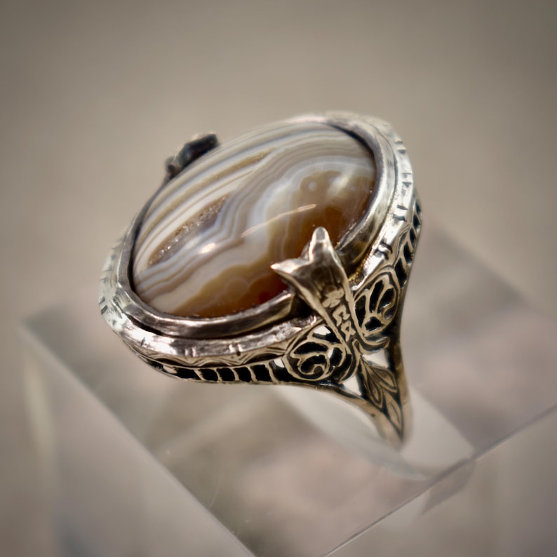 Puede incluir: Un anillo de plata con una piedra preciosa de &aacute;gata ovalada y bandeada, con capas conc&eacute;ntricas de color marr&oacute;n, blanco y bronceado. La banda de metal muestra detalles intrincados de filigrana calada en los lados, enmarcando la piedra pulida.