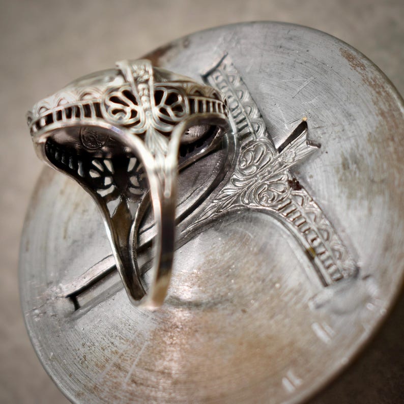 Puede incluir: Un anillo de plata con una intrincada galer&iacute;a de filigrana y una banda lisa y pulida. El trabajo en metal muestra patrones calados detallados en los lados, que sostienen un engaste central que descansa sobre una superficie met&aacute;lica circular.