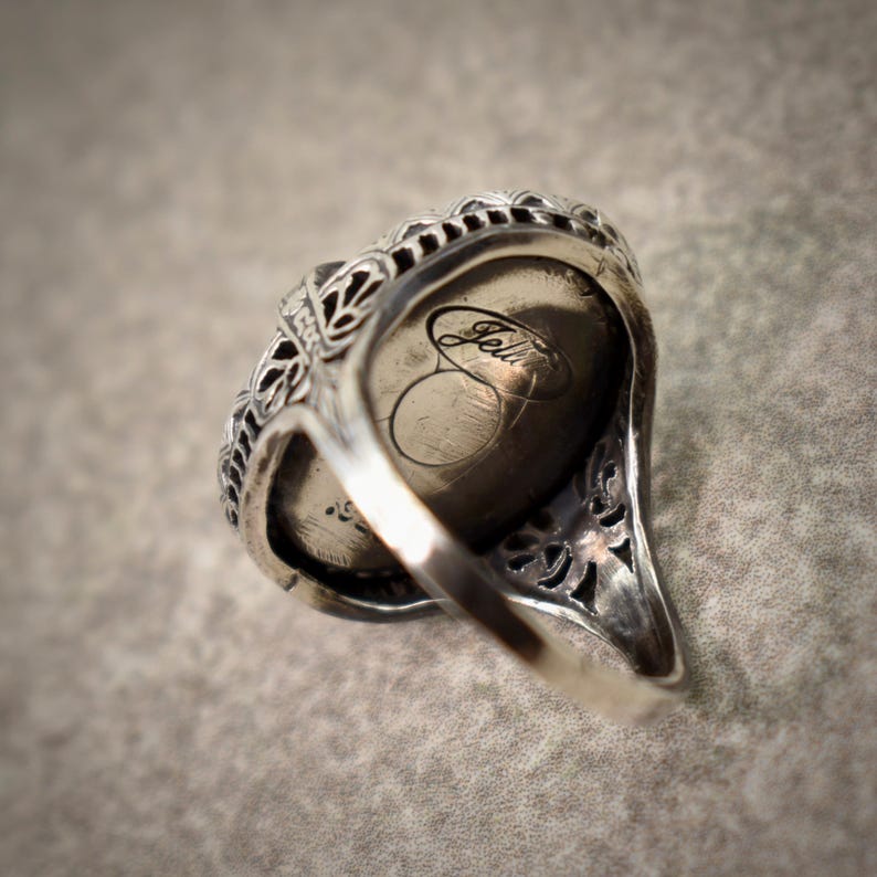Puede incluir: Un anillo de plata con una banda de filigrana intrincada y una piedra central ovalada, lisa y oscura. La parte inferior del anillo muestra una marca de firma grabada clara que dice "Jellin" en letra cursiva, visible dentro del engaste met&aacute;lico.