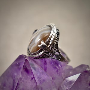 Puede incluir: Un anillo de plata con una piedra preciosa de &aacute;gata ovalada de bandas marrones y blancas. La banda de metal muestra intrincados detalles de filigrana calada en los laterales, exhibiendo un patr&oacute;n decorativo que sostiene la montura central.