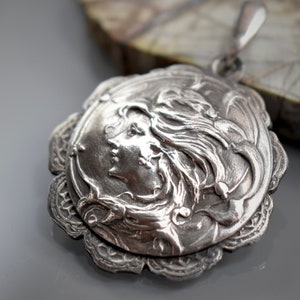 May include: A silver pendant featuring a detailed, embossed design of a woman's face with flowing hair. The pendant has a decorative, scalloped edge.
