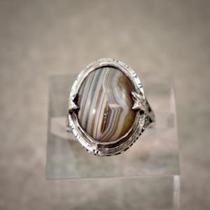 Puede incluir: Un anillo de plata con una piedra preciosa de &aacute;gata ovalada con bandas de colores marr&oacute;n, blanco y gris. La banda de metal muestra patrones intrincados en relieve en los lados, y la piedra est&aacute; asegurada por cuatro garras met&aacute;licas puntiagudas.