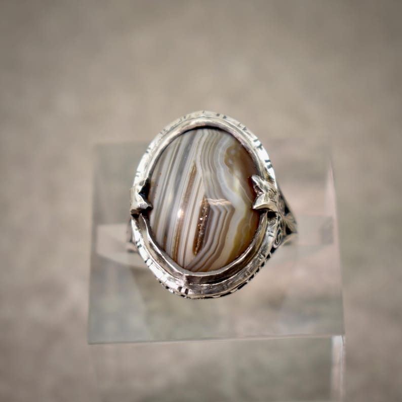 Puede incluir: Un anillo de plata con una piedra preciosa de &aacute;gata ovalada con bandas de colores marr&oacute;n, blanco y gris. La banda de metal muestra patrones intrincados en relieve en los lados, y la piedra est&aacute; asegurada por cuatro garras met&aacute;licas puntiagudas.