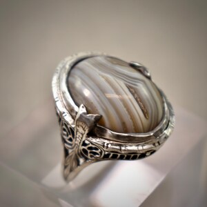Puede incluir: Un anillo de plata con una piedra preciosa de &aacute;gata pulida de forma ovalada con patrones de bandas marrones y blancas. La banda de metal muestra un intrincado trabajo de filigrana en relieve en los lados, enmarcando la piedra central.