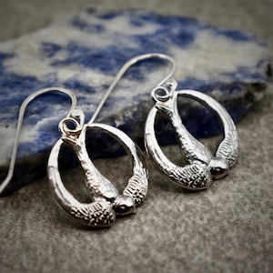 May include: A pair of silver earrings with a unique design. Each earring features a curved, oval shape with textured detailing and a small, round bead at the bottom. The earrings have a hook closure.