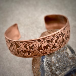May include: A copper cuff bracelet featuring an intricate, embossed scrollwork pattern across its entire outer surface. The metal has a warm, reddish-brown tone with darker recessed areas that highlight the detailed floral and leaf-like engravings.