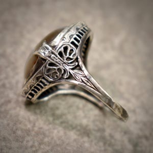 Puede incluir: Un anillo de plata con una piedra ovalada, lisa y de color marr&oacute;n claro engastada en una montura de metal decorativa. La banda y el engaste muestran intrincados patrones grabados y detalles de filigrana calada en los laterales.